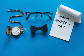Happy Fathers Day Concept. Sheet of paper with congratulations. Transparent glasses, black stylish paper bow tie, vintage pocket watch and notebook on blue background. Copy space.