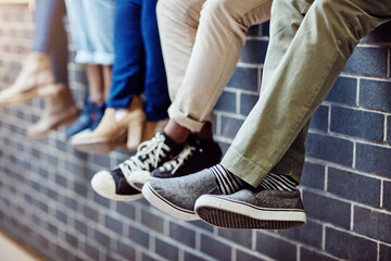 Brick wall, student feet and friends outdoor on university campus together with sneakers. Relax, urban youth and foot at college with people legs ready for education, study and shoes while sitting - Powered by Adobe