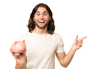 Young handsome man holding a piggybank over isolated background surprised and pointing finger to...