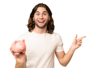 Young handsome man holding a piggybank over isolated background surprised and pointing finger to the side