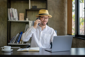 Asian businessman sitting at homeoffice front of laptop. talking with his crew via smartphone while checking data on laptop. 
