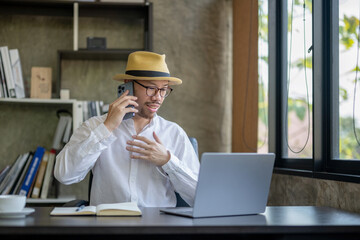 Asian businessman sitting at homeoffice front of laptop. talking with his crew via smartphone while checking data on laptop.