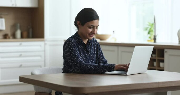 Millennial Indian woman sitting in kitchen and working on laptop, smile while typing message on notebook keyboard, share message, doing freelance telecommute work from home using internet connection