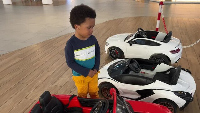 Frustrated 3-year-old balck toddler fustrated and impatient to ride a toy car inside a mall. We wears curly hair and a navy blue colot shirt.