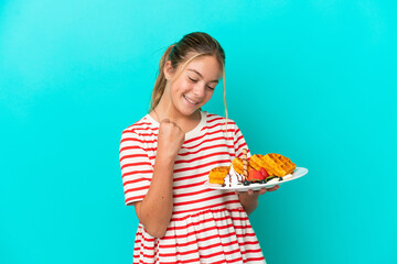 Little caucasian girl holding waffles isolated on blue background celebrating a victory