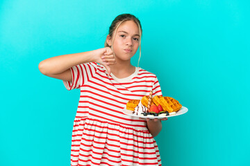 Little caucasian girl holding waffles isolated on blue background showing thumb down with negative expression