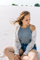 a girl in a grayT-shirt and denim shorts in a white cape, a cardigan, a poncho with a hat on her head in the desert, on the beach on the sand