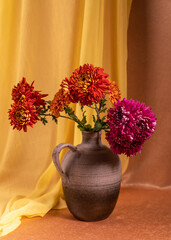 A bouquet of red, pink dahlias in a vase on an orange background, next to it lies a yellow scarf. Still life, postcard