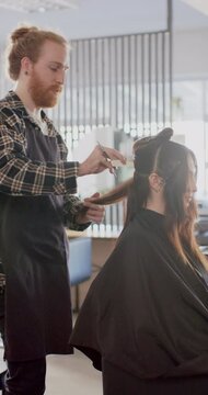 Vertical video of caucasian male hairdresser cutting client's hair at hair salon, in slow motion
