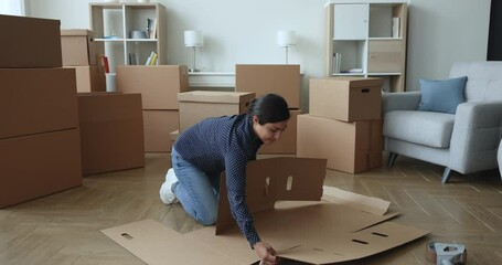 Young single Indian female assembling parts of big cardboard box for personal stuff storing at relocation day, leaves to her new own or rented house, prepare dwelling for remodeling or removal work