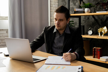 Fototapeta premium A young man in a business suit is working on a laptop while sitting in the office. A man in a black jacket sits in front of a laptop and fills out paperwork.