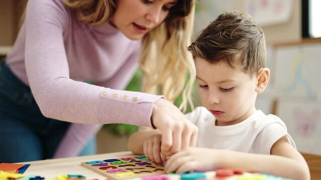Woman and boy playing with maths puzzle game sitting on table at kindergarten