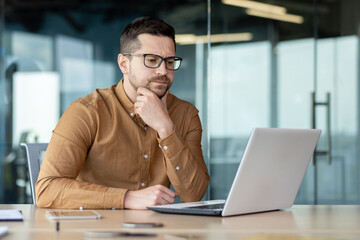 Confused young male businessman sits in the office in front of a laptop and looks thoughtfully at the screen, concentrates on a new project, solves a problem