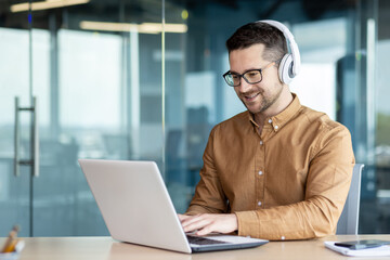 Portrait of a young smiling male student sitting in the office wearing headphones and studying...