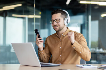 A young man, an office worker, a freelancer works in the office with a laptop and listens to music in headphones, rests, took a break, moves his hands, dances