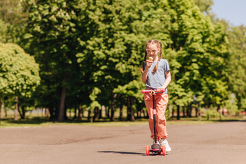 A child riding a scooter. Active outdoor activities for children. Summer sports for preschool children. A little girl in a spring park. The girl eats ice cream and enjoys