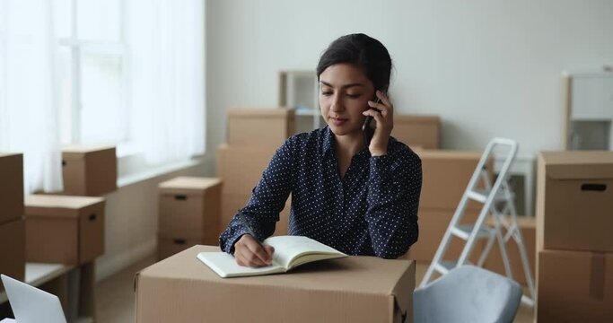 Young Indian businesswoman, female small business owner, talks to client on cellphone, jotting information, take notes, working surrounded by heap of carton boxes ready for sending to customers. SME