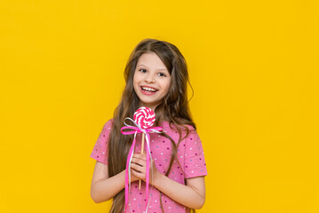 A happy little girl holds a caramel lollipop in her hands and smiles, a girl with a candy on an isolated yellow background.