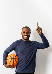 A dark-skinned man poses on a white background in casual clothes with a basketball. The guy points up with his finger,promote party,ad,copyspace vertically.