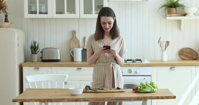 Happy young adult girl using smartphone in kitchen, typing, consulting cook blog, reading online recipe, preparing organic healthy salad for dinner, cutting fresh vegetables, taking picture of table