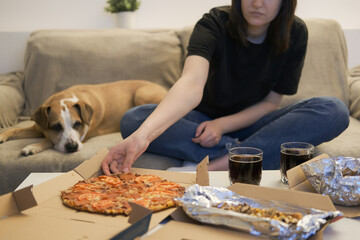 Eating delivered pizza at home. Woman grabbing a slice of pizza, dog on the couch watching, focus on food