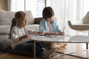 Focused little brother and sister doing primary school homework at home, learning to write, drawing in paper albums, scratching doodles, sitting on warm heating floor