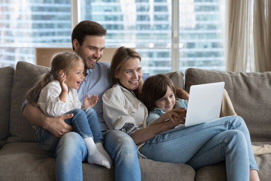 Happy couple of parents using online learning app on laptop, for educating little kids, resting on couch together, holding digital device, enjoying wireless technology, domestic internet connection