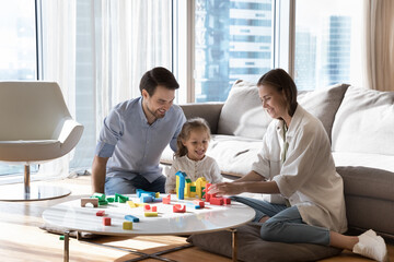 Cheerful family couple and little kid girl stacking wooden colorful toy blocks at home, constructing castle, tower, house, playing smart game, enjoying activity for child development