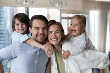 Cheerful young parents piggy backing little kids, holding children, looking at camera with toothy smile, promoting white healthy teeth, dental care, dentistry service