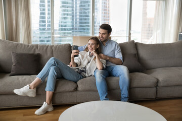 Happy young couple using mobile phone together, resting on big soft comfortable sofa in city apartment, making video call, using ecommerce application for Internet communication