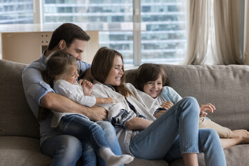 Cheerful young parents playing with little children on couch at home, enjoying family leisure, cuddling, tickling kids, giggling, laughing, having fun, feeling happiness, bonding, love