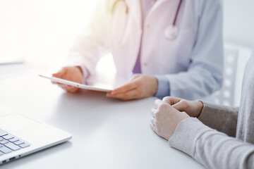 Doctor and patient sitting at the table in clinic. The focus is on female physician's hands using tablet computer, close up. Medicine and healthcare concept