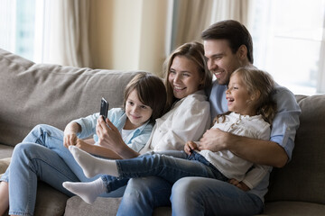 Young mother and father hugging cheerful kids on home sofa, taking happy family picture on smartphone, using mobile phone together, for online communication, enjoying domestic Internet technology