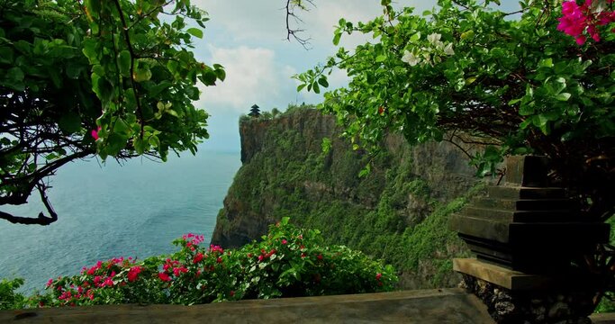 Ocean view and ancient religious monument on egde of cliff Uluwatu Temple on Bali Island. Indonesia. Flamboyant pink flowers.