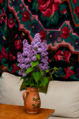 Syringa vulgaris, lilac flower, blooming branches in clay jug on table with traditional carpet background, in country house