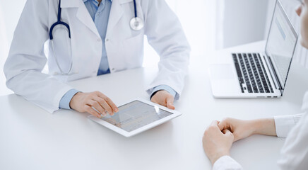 Fototapeta premium Doctor and patient sitting near each other at the desk in clinic. The focus is on female physician's hands pointing into tablet computer touchpad, close up. Medicine concept
