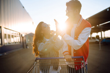 Stylish man and woman having fun and riding shopping cart. Black friday. Consumerism, sale, discounts, lifestyle concept.