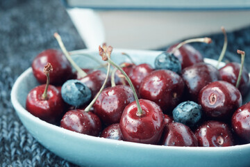 Bowl full of ripe red cherries and books on a dark blue knitted blanket.