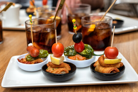 Tapa de pinchos t&iacute;picos de la gastronom&iacute;a espa&ntilde;ola, acompa&ntilde;ados de unos vasos con vermut de fondo