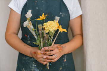 Close-up of young artist holding bouquet made of flowers and paint brushes in studio