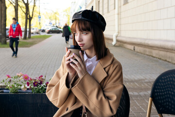 A lovely young brunette with a bob haircut, wearing a beret and a beige classic coat, is sitting on a cafe terrace with a coffee, pastries and a newspaper. Outdoor morning, spring.