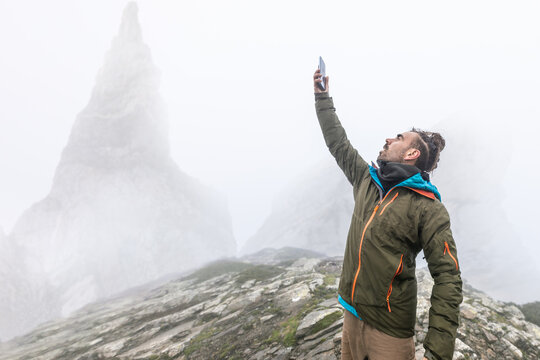 man on the mountain in the fog with rain, wet, long hair looking for coverage with cell phone, wild scene, looking for wifi concession in nature, high mountains in the background