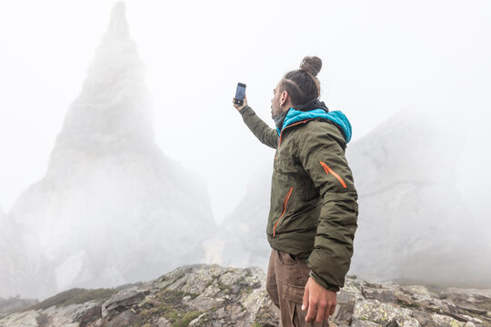 man on the mountain in the fog with rain, wet, long hair looking for cover with cell phone, wild scene, taking pictures in nature, high mountains in the background