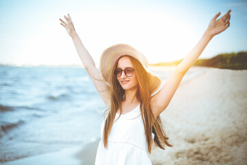 Fototapeta na wymiar Portrait of a happy smiling woman in free happiness bliss on ocean beach standing with a hat and sunglasses. A female model in a white summer dress enjoying nature during travel holidays vacation