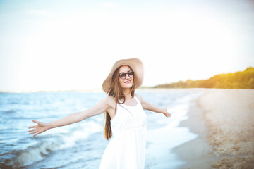 Fototapeta na wymiar Portrait of a happy smiling woman in free happiness bliss on ocean beach standing with a hat and sunglasses. A female model in a white summer dress enjoying nature during travel holidays vacation