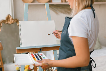 Close-up of woman standing at an easel with blank paper mockup painting with watercolour paints.