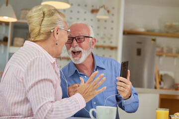 Happy excited senior couple sharing earphones listening to the music while sitting at home