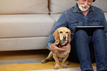 Close up of senior man sits on the floor with his dog and using tablet pc. Elderly man listening to music, podcast, watch movie, video call with family or friends. Leisure and retirement concept