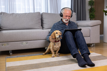 Senior man sits on the floor with his dog and using tablet pc. Elderly man with headphones listening to music, podcast, watch movie, video call with family or friends. Leisure and retirement concept - 602110969