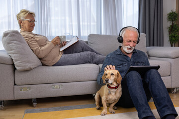 Elderly couple spending leisure time at home with their dog. Spouses reading book, listening to music, podcast or watching a movie while sitting on sofa. Happy retirement concept.